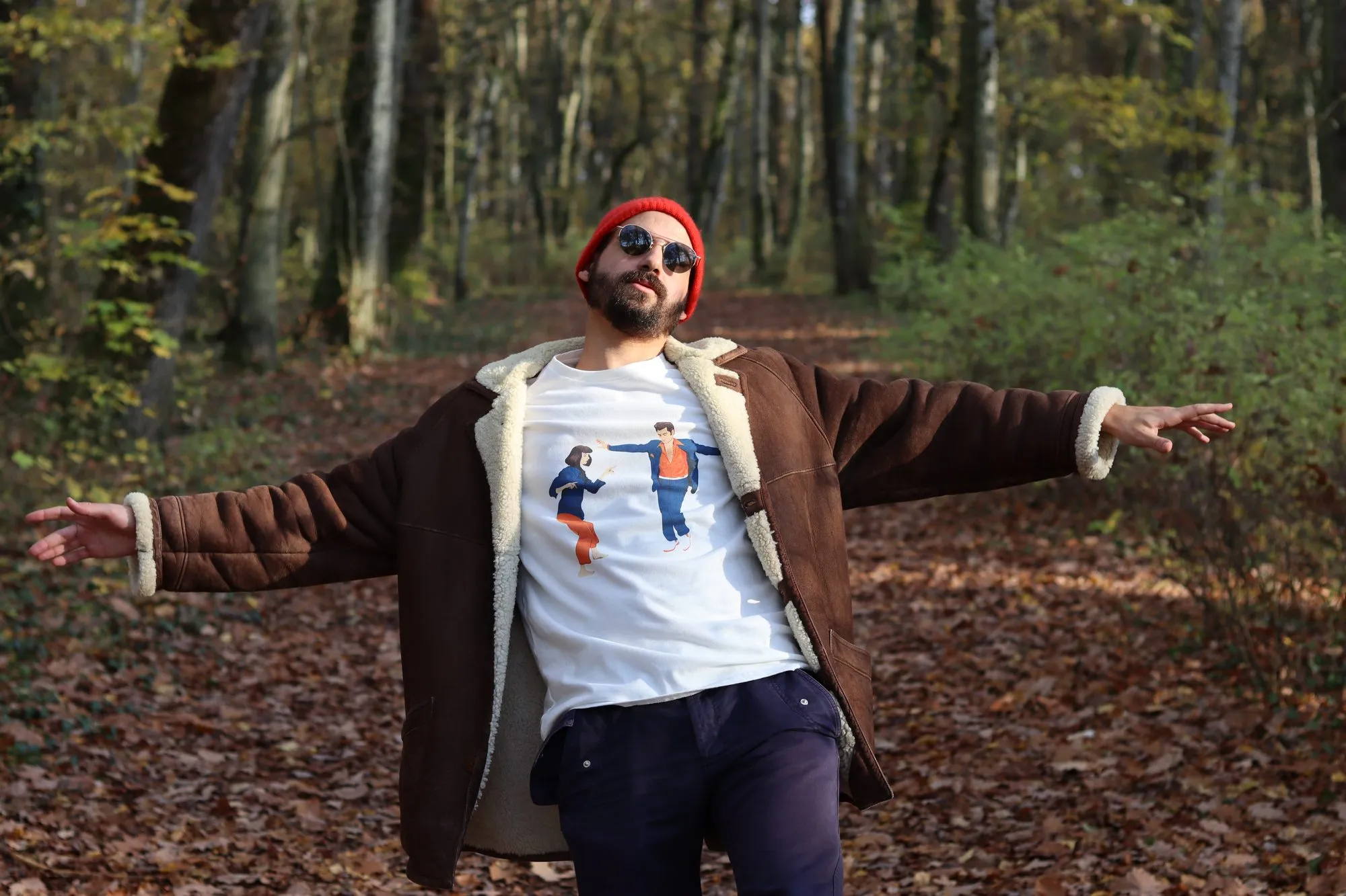 Guillaume, co-founder of WAAS, arms wide open in an autumn forest, in a red beanie, sunglasses, a sheepskin coat and The Lovers tee